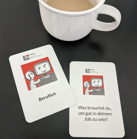 Foto: Zwei Spielkarten liegen auf schwarzer Fläche. Im Hintergrund steht eine Kaffeetasse.